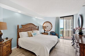Quarto com uma cama grande, cabeceira de madeira, mesa de cabeceira com luminária, cômoda e vista para um prédio alto através de uma porta de vidro.