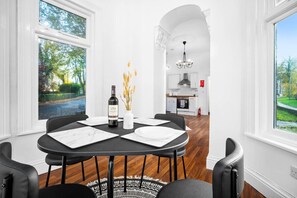 A dining area with a table set for four, a bottle of wine, and a vase with dried flowers. The room has white walls, wooden flooring, and large windows with a view of greenery outside.