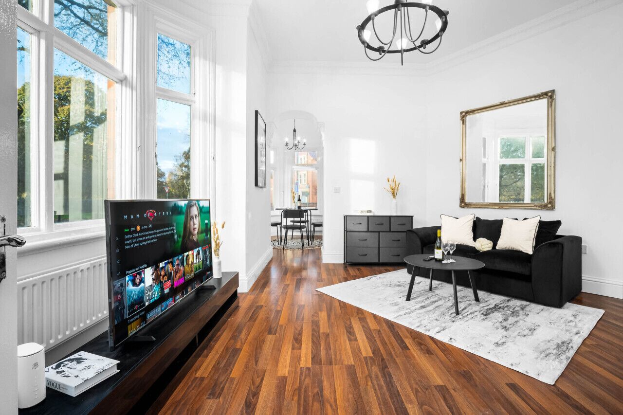 A modern living room with a large flat-screen TV, a black sofa, a coffee table, and a chandelier.