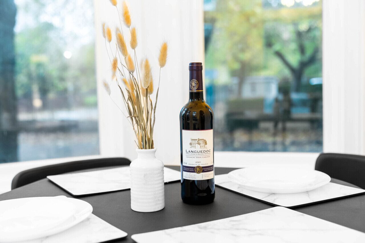 Una botella de vino de Languedoc sobre una mesa con platos blancos y un jarrón con plantas secas.