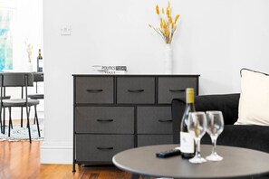 A modern living room with a grey cabinet, a black sofa, a round table with wine glasses and a remote control, and a vase with dried flowers on the wall.