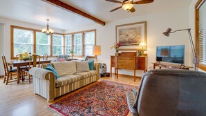 A spacious living room with a large window, a ceiling fan, a television, and a dining area with a table and chairs. - Historic District Home - Walk to Willapa Bay! (Oysterville)
