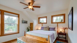 A bedroom with a wooden bed, a ceiling fan, two windows, a bedside table with a lamp, and a dresser. - Historic District Home - Walk to Willapa Bay! (Oysterville)