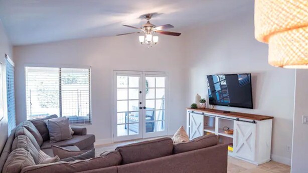 A living room with a brown sectional sofa, a ceiling fan, a TV on a wooden stand, and a sliding glass door leading to an outdoor area. - Perfectly-Located, Charming Family Home w/ Pool (Phoenix)