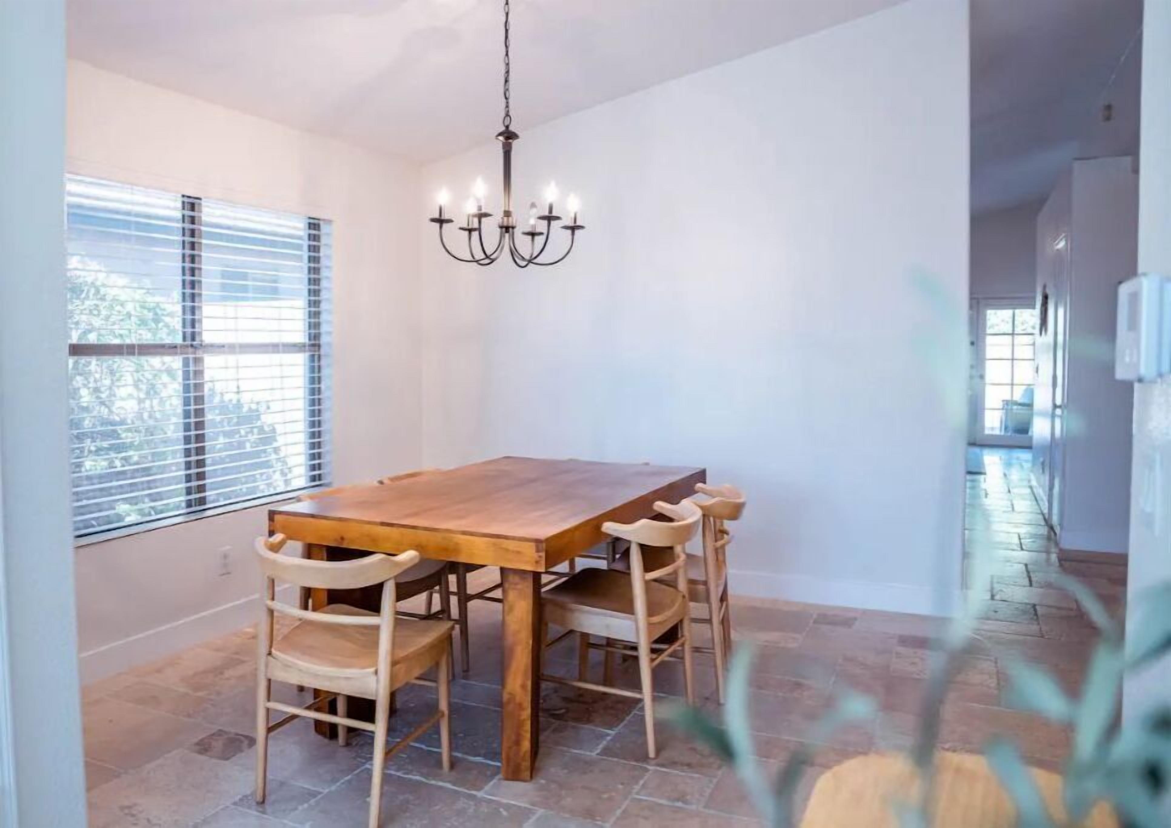 A dining area with a wooden table and chairs, a chandelier, and a view through a window with blinds.