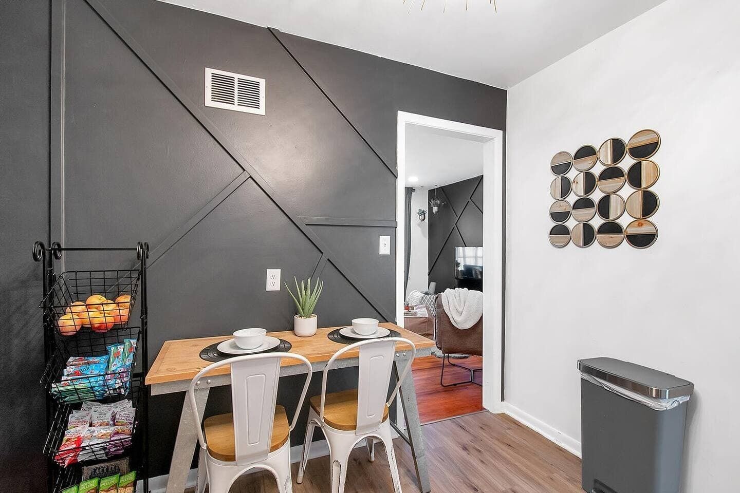 Un comedor moderno con una mesa de madera, sillas blancas y un estante metálico negro con aperitivos.