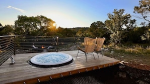 A wooden deck with a hot tub and wicker chairs overlooking a body of water, surrounded by trees.