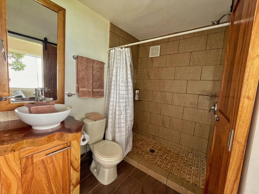 A bathroom with a wooden vanity, a white sink, a toilet, and a shower with a beige tiled wall.