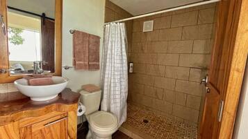 A bathroom with a wooden vanity, a white sink, a toilet, and a shower with a beige tiled wall.