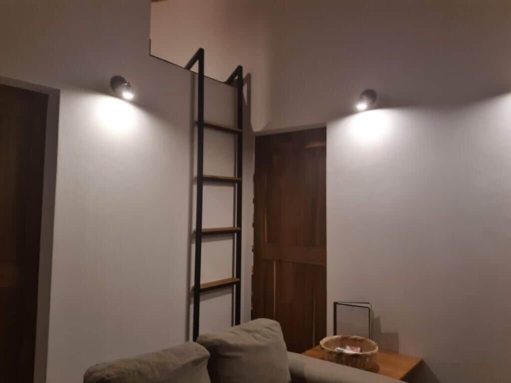 Una habitación con sofá, una escalera de madera y dos luces fijadas a la pared.