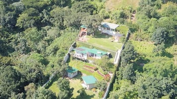 An aerial view of a property with multiple buildings surrounded by dense forest. There are several structures with green roofs, a visible swimming pool, and a paved pathway connecting the buildings.