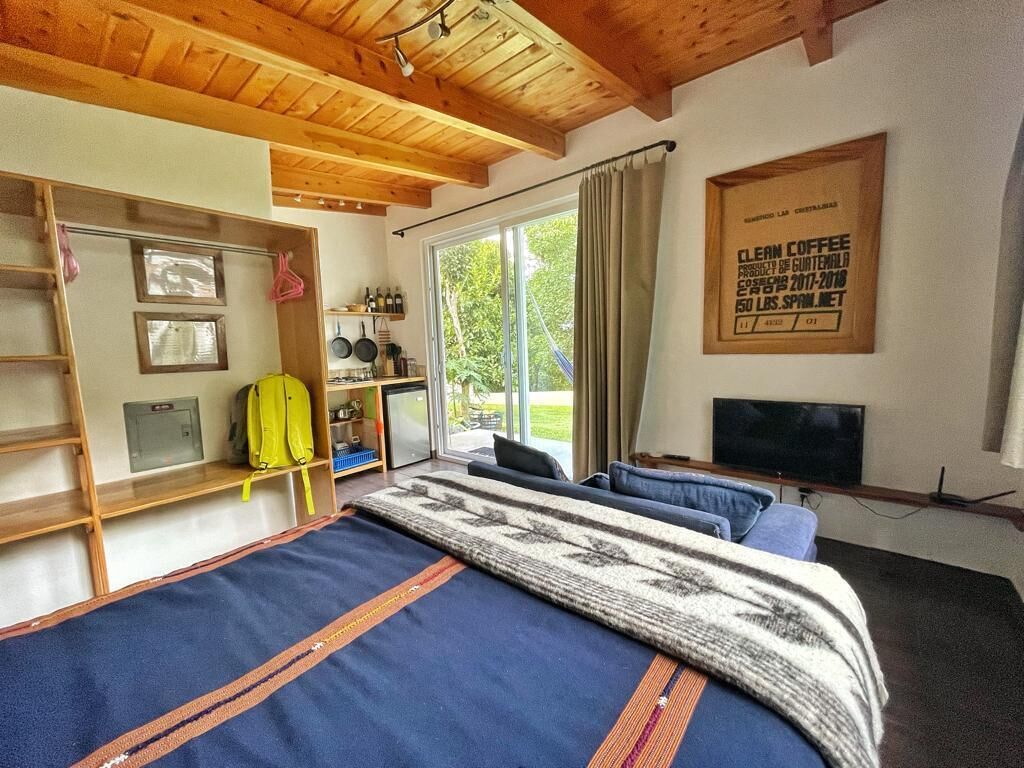 A bedroom with a bed, a TV, a window with curtains, a shelf with bottles, and a framed certificate on the wall.