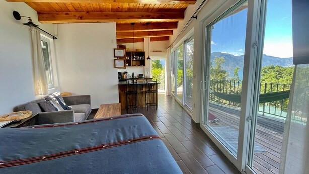 Una habitación moderna con una cama grande, un rincón de descanso y una cocina. Destaca por su techo de madera y amplias ventanas con vistas a la montaña.