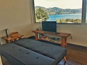A room with a large window, a television on a wooden stand, a small table with a basket, and a cushioned bench. - Refugio del Volcan (San Pedro La Laguna)