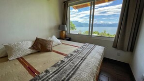 Un dormitorio con una cama grande, una mesita de noche con lámpara y ventana con vistas a un lago y montañas.