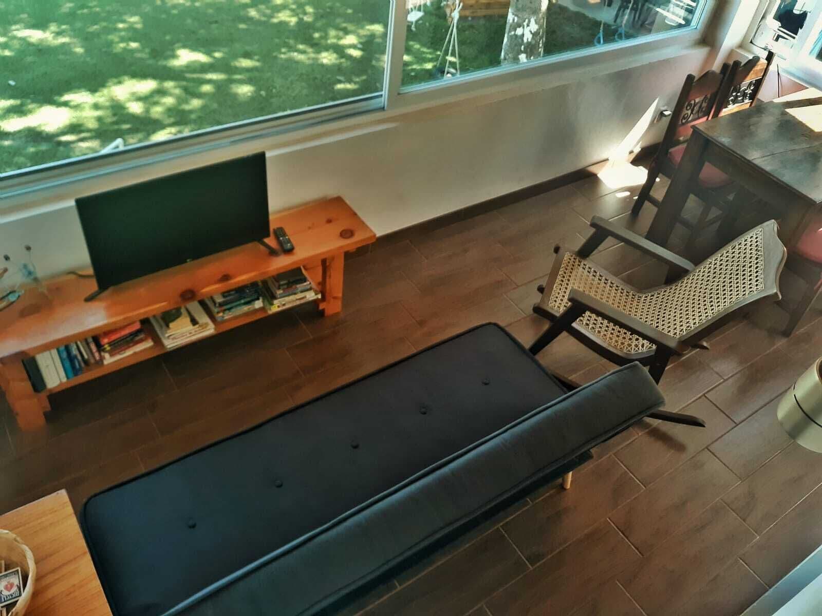A modern living room with a large window, a flat-screen TV, a wooden entertainment unit, a black sofa, and a wicker chair.