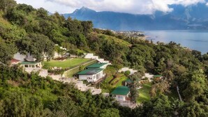 Una propiedad en una ladera con varias construcciones, incluyendo una casa con techo verde, rodeada de vegetación exuberante y vistas a un lago y montañas.