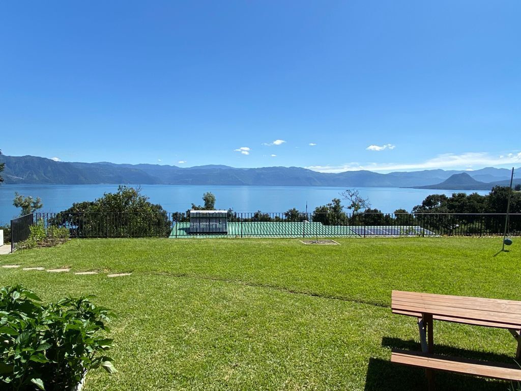 A grassy area with a bench, a fenced sports court, and a view of a lake with mountains in the distance.