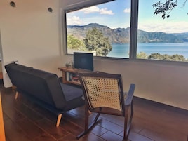 Habitación con un ventanal grande que ofrece vistas a un lago y montañas. Hay un televisor sobre un soporte de madera, un sofá y una silla con respaldo tejido.