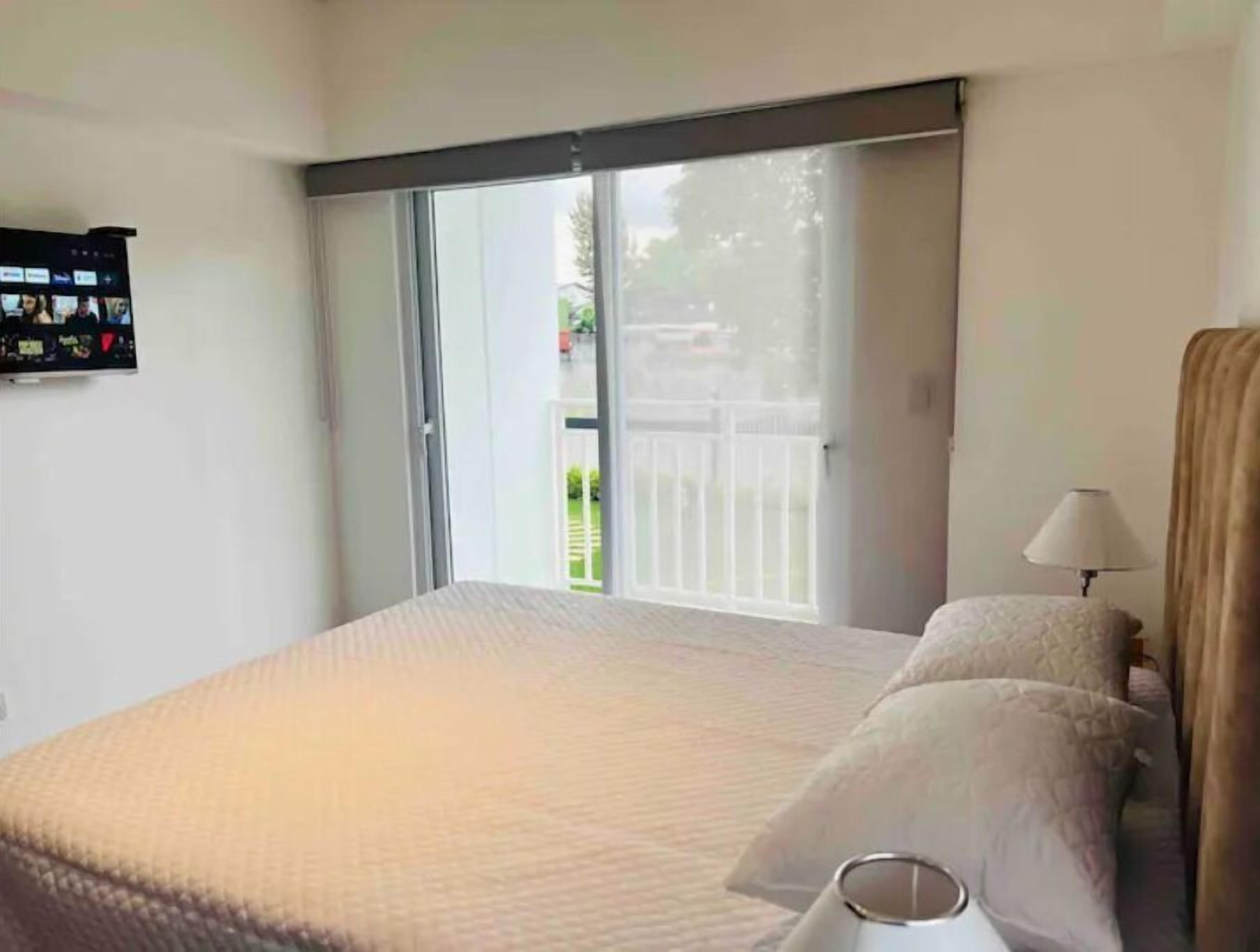 A bedroom with a large bed, a TV mounted on the wall, and a sliding glass door leading to a balcony.