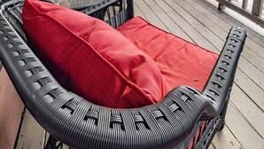 Een overdekt terras met rieten meubels, rode kussens en een potplant.