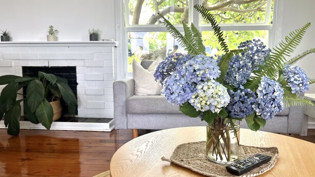Ein Wohnzimmer mit grauem Sofa, einem weißen Kamin, einem runden Couchtisch mit Fernbedienung und einer Vase mit blauen Hortensien.