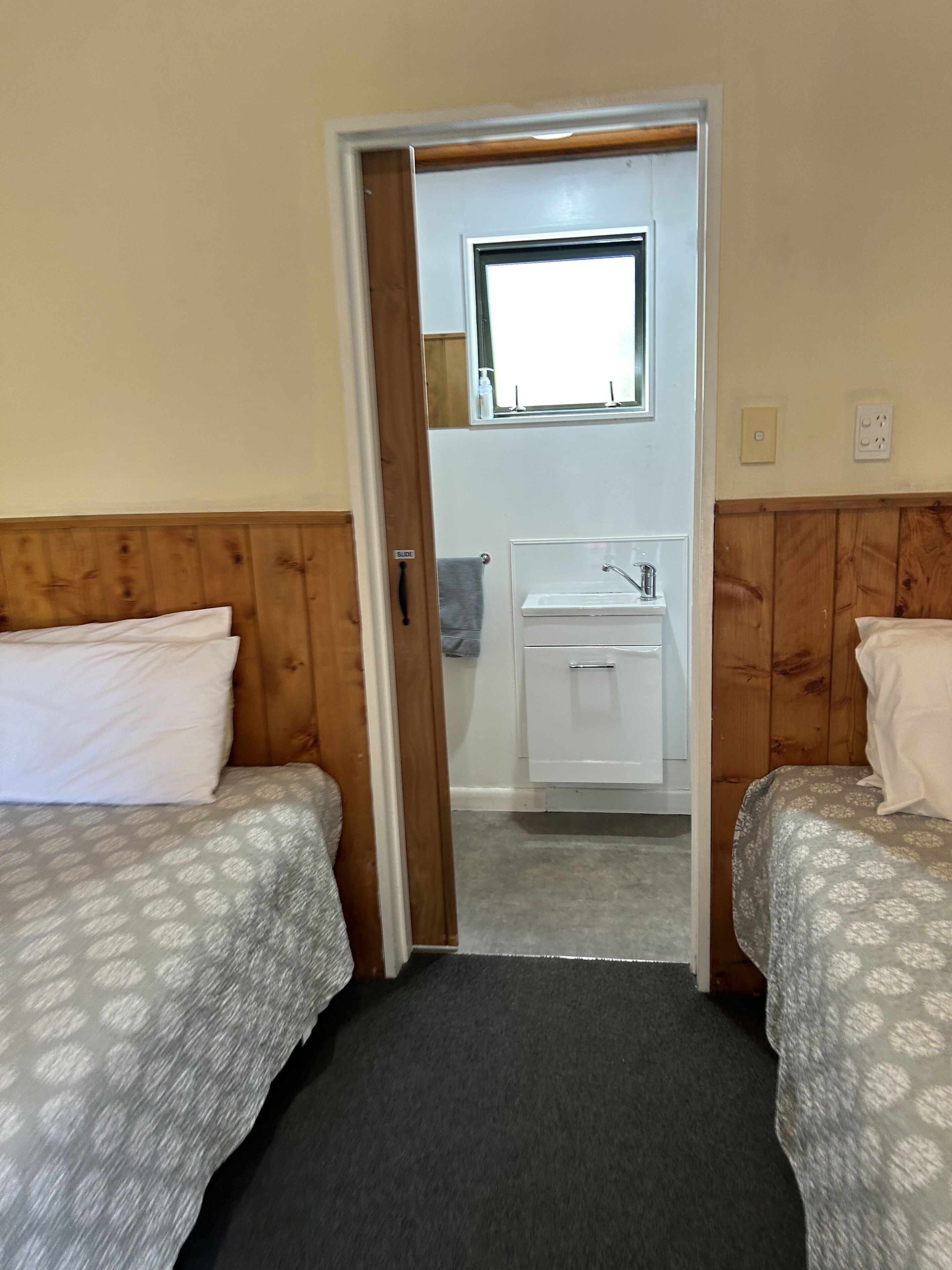Una habitación con dos camas, un baño pequeño con lavamanos y una ventana sobre el espejo.