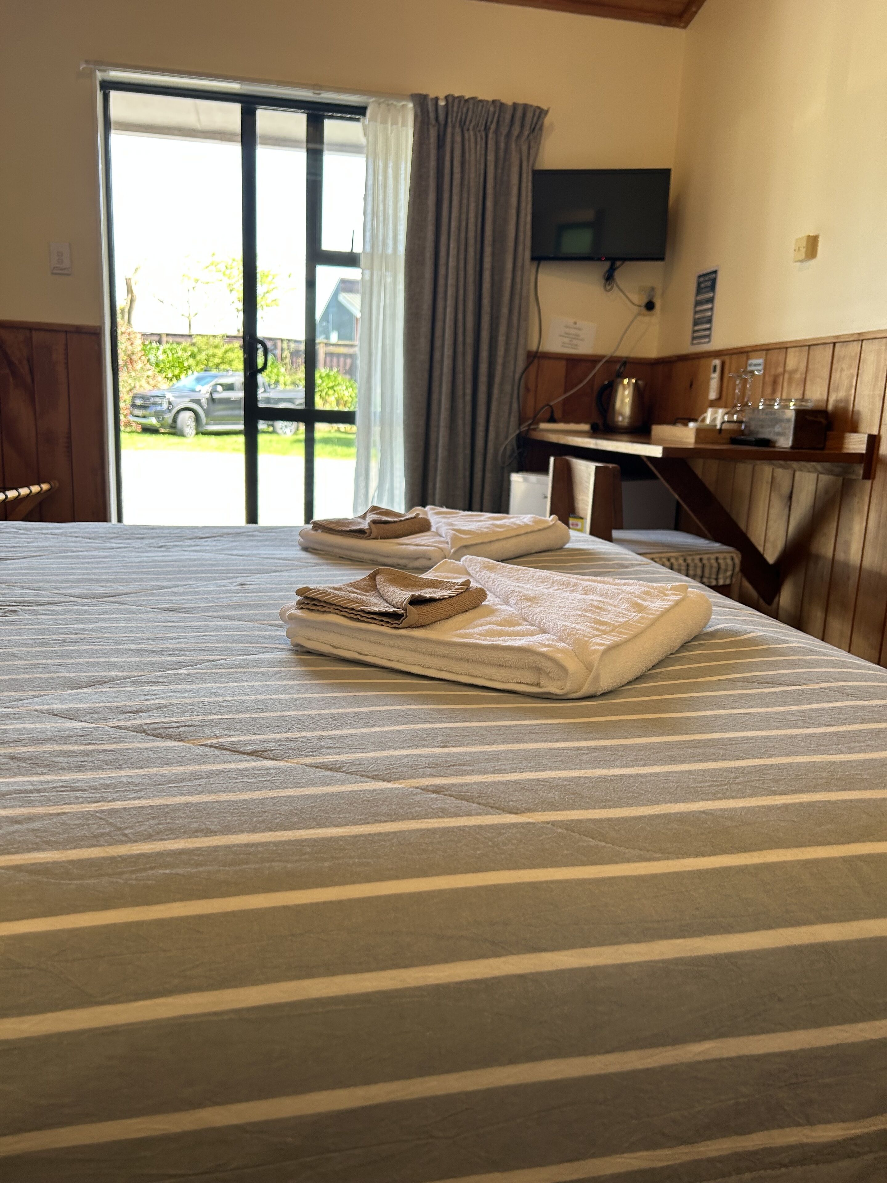 Quarto de hotel com cama, televisão fixada na parede e vista para o exterior por uma grande janela.