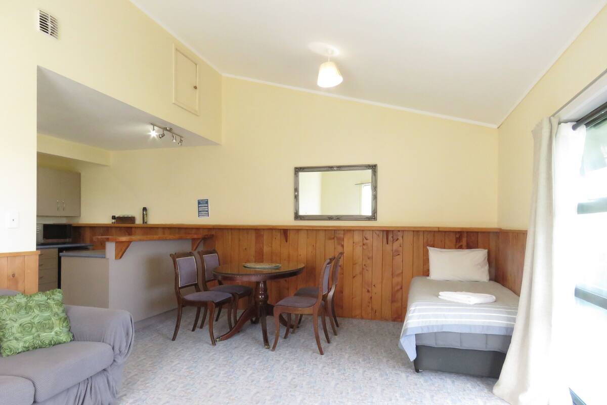 Une pièce compacte comprenant un coin cuisine, un espace repas avec une table et des chaises, un lit avec un oreiller et un miroir au mur.