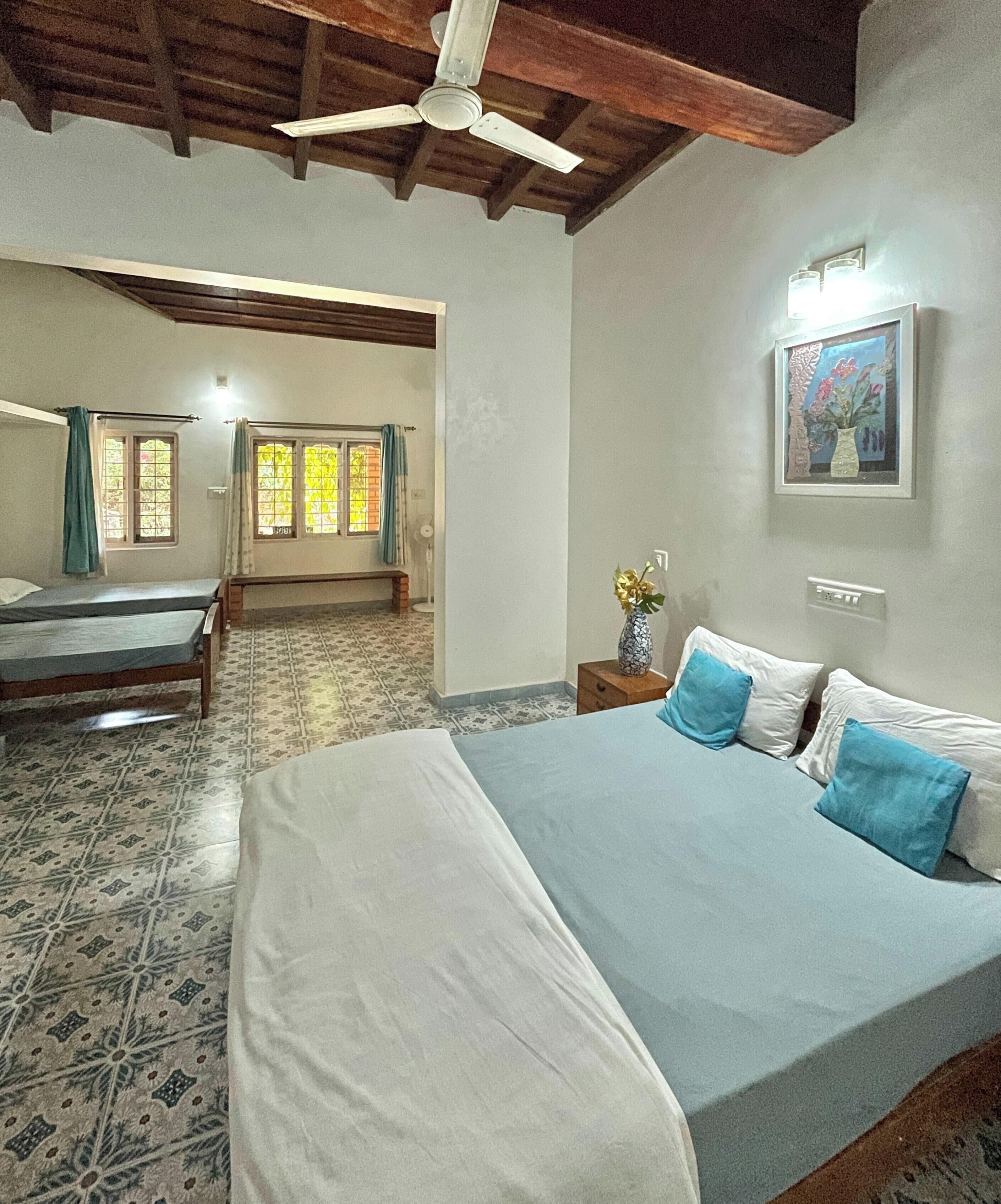 A bedroom with a large bed, a smaller bed, a ceiling fan, a window with curtains, and a wall-mounted light fixture.