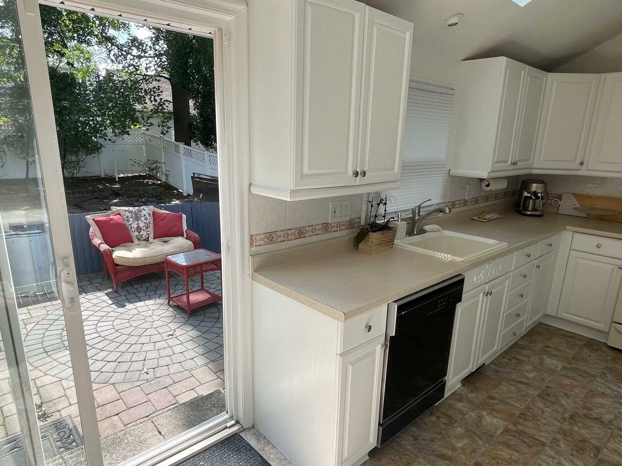 A kitchen with white cabinets, a dishwasher, and a sink. There is a small outdoor seating area with a red chair and a table.