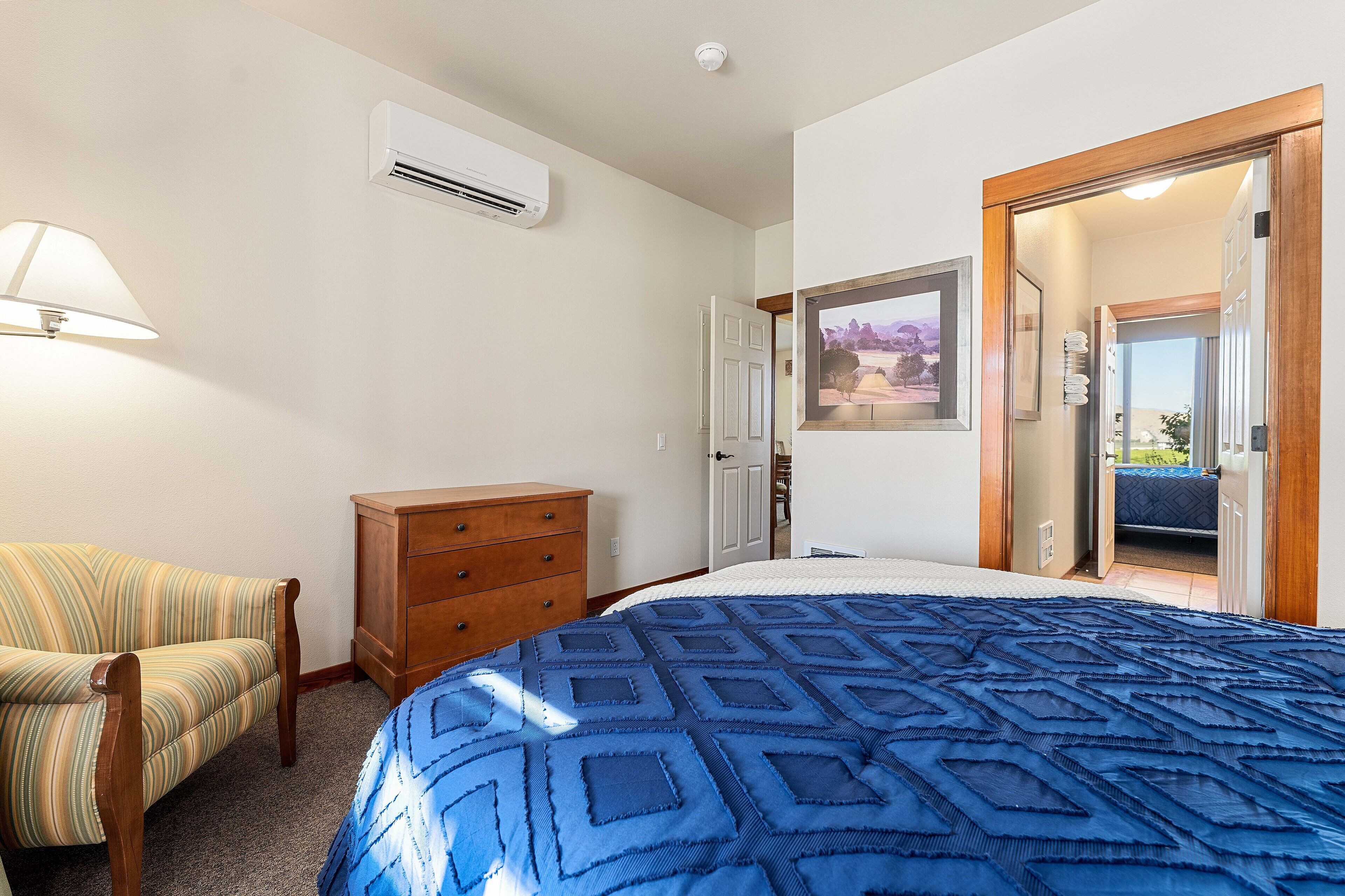 Une chambre à coucher avec un grand lit, une commode en bois, un fauteuil à rayures et une porte donnant sur une autre pièce.