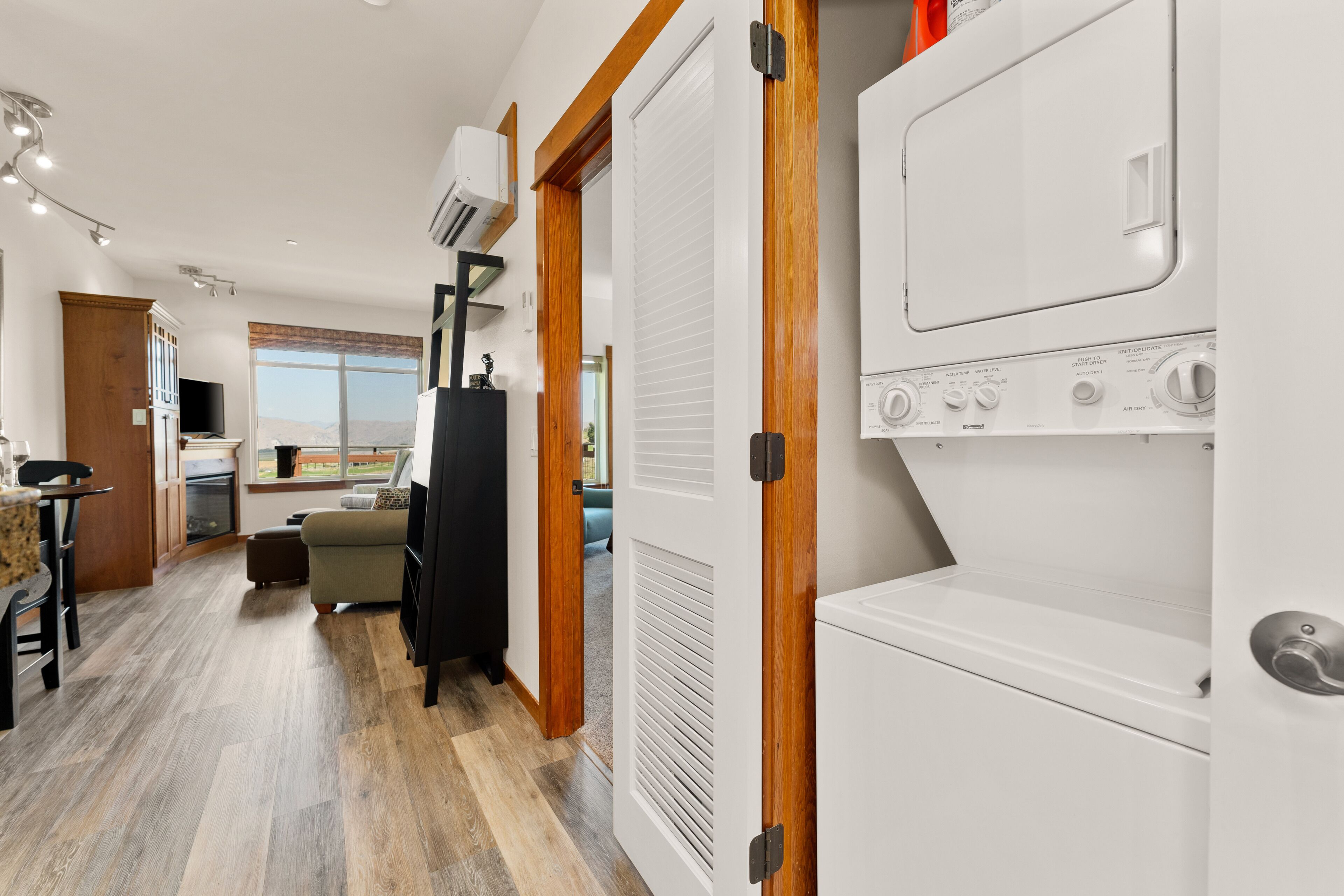 A modern kitchen with a built-in washer and dryer, wooden flooring, and a large window offering a view of the outdoors.