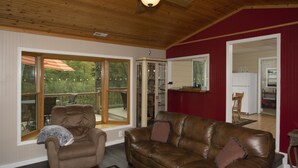 A living room with a brown leather couch, a recliner, and a ceiling fan. There are large windows and a view of the outdoors. - Quiet Cabin Setting on small river
 (Chase)