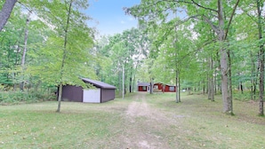 A forested area with a dirt path leading to a small building with a red roof. - Quiet Cabin Setting on small river
 (Chase)