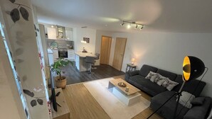 A modern living room with a sofa, coffee table, and a lamp. There is a kitchen area in the background with a dining table and chairs. - New comfort apartment. Basement, good for excursions, Europapark, Strasbourg (Ortenberg)