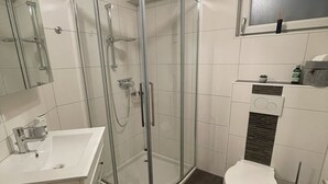 A modern bathroom with a glass shower enclosure, a white sink, and a toilet. - New comfort apartment. Basement, good for excursions, Europapark, Strasbourg (Ortenberg)