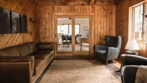 A cozy cabin interior with wooden walls, a brown leather sofa, a grey armchair, and a small table with a lamp. - Hemlock Haven,Hot-tub,Private,30 min to Penn State (Mill Hall)