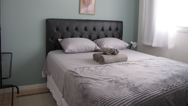 Een slaapkamer met een gestoffeerd hoofdeinde, twee kussens, een opgerolde handdoek en een ingelijst beeld aan de muur.