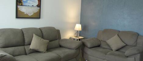 A living room with two grey sofas, a wooden coffee table with a plant, a lamp on a side table, and a painting of a beach scene on the wall.