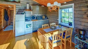 Une cuisine chaleureuse, aménagée dans le style d’un chalet, avec un plancher en bois, une table à manger avec des chaises, un réfrigérateur, un four, un évier et une fenêtre donnant sur un paysage verdoyant.