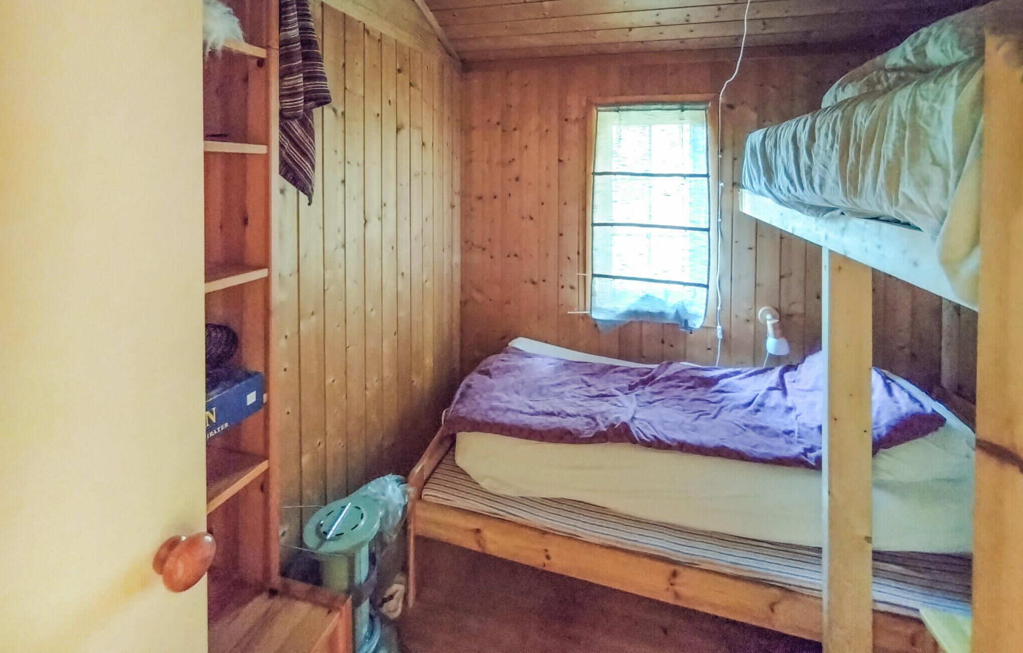 A small, wooden cabin room with a bunk bed, a window with curtains, and a shelf with a water bottle and a box.