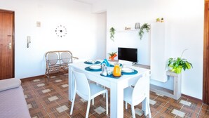 Un comedor con una mesa blanca preparada para cuatro personas, un televisor en la pared y un reloj.