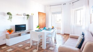 Un salón luminoso con un sofá blanco, una mesa de comedor con platos y un televisor sobre un soporte de madera.