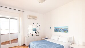 Un dormitorio con cama, mesita de noche, cómoda, espejo, aire acondicionado de pared y ventana con vista a barcos.