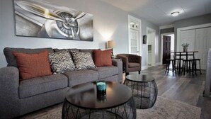 Ein Wohnzimmer mit grauem Sofa, gemusterten Kissen, zwei runden Tischen und einem großen abstrakten Gemälde an der Wand.