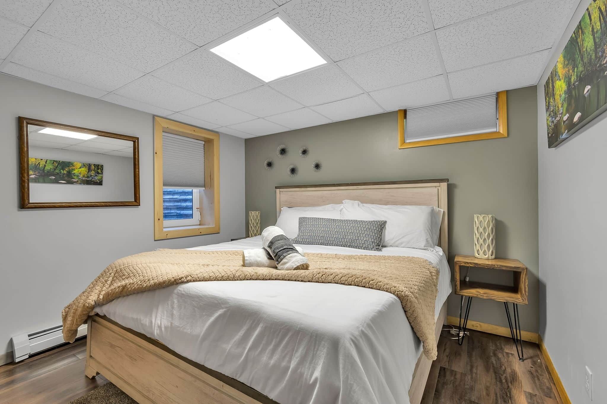 A neatly made bed with a white comforter and a beige blanket, a bedside table with a decorative vase, and a wall-mounted mirror.