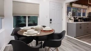 Een eetgedeelte met een ronde tafel voor vier personen, zwarte stoelen, een witte deur en een keuken met kasten en een aanrecht.