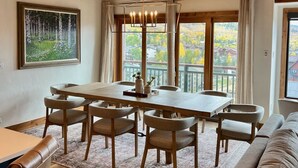 Un comedor con una mesa y sillas de madera, una araña y un cuadro de un bosque.