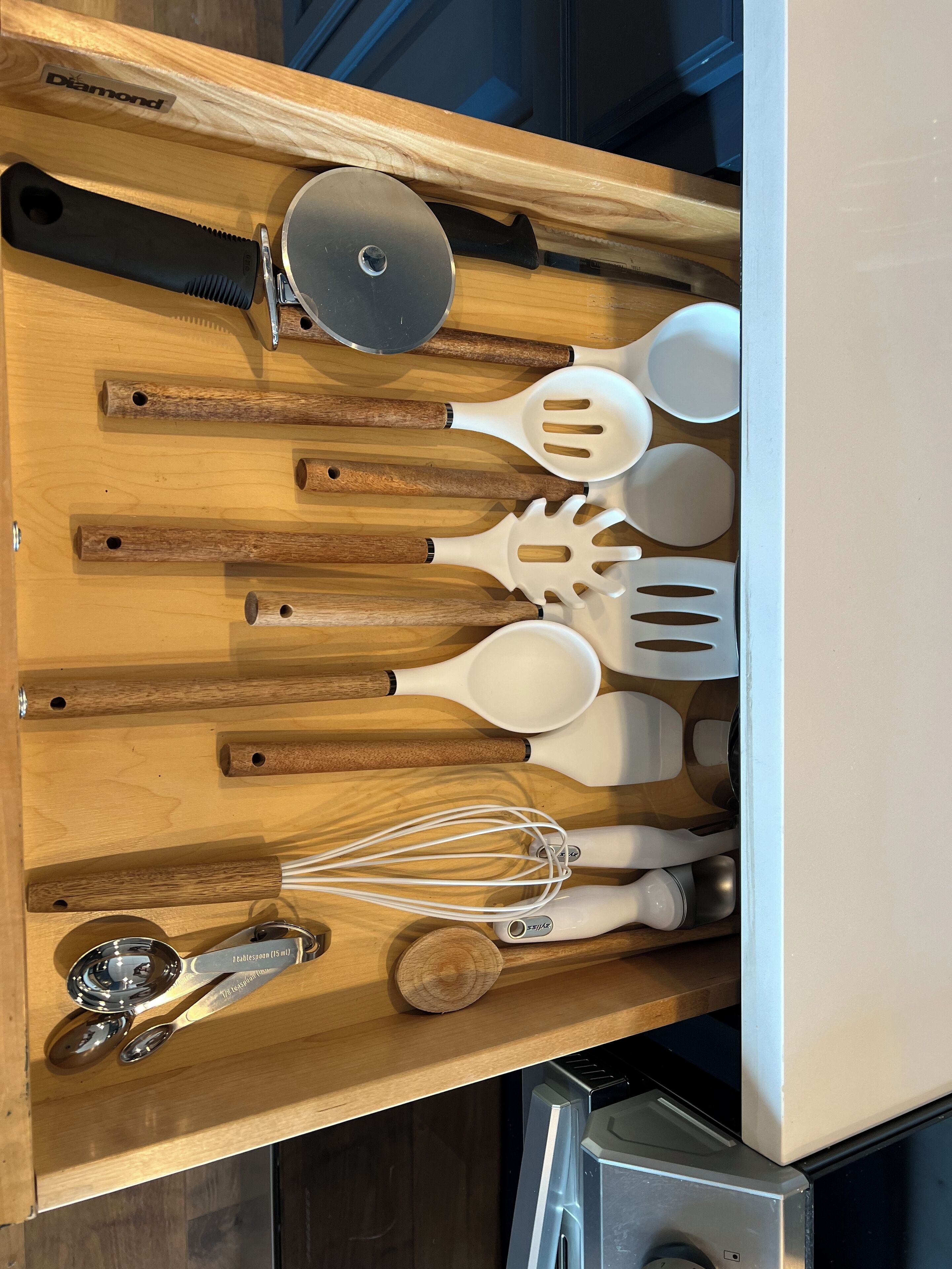 A kitchen drawer with wooden handles, a metal lid, white plastic spatulas, a whisk, and a pair of scissors.